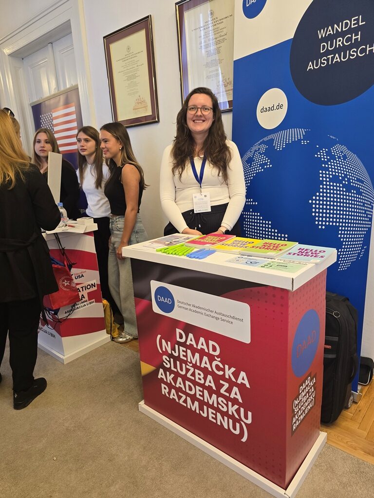 A smiling representative stands behind the DAAD table in front of a blue banner reading “Wandel durch Austausch”.
