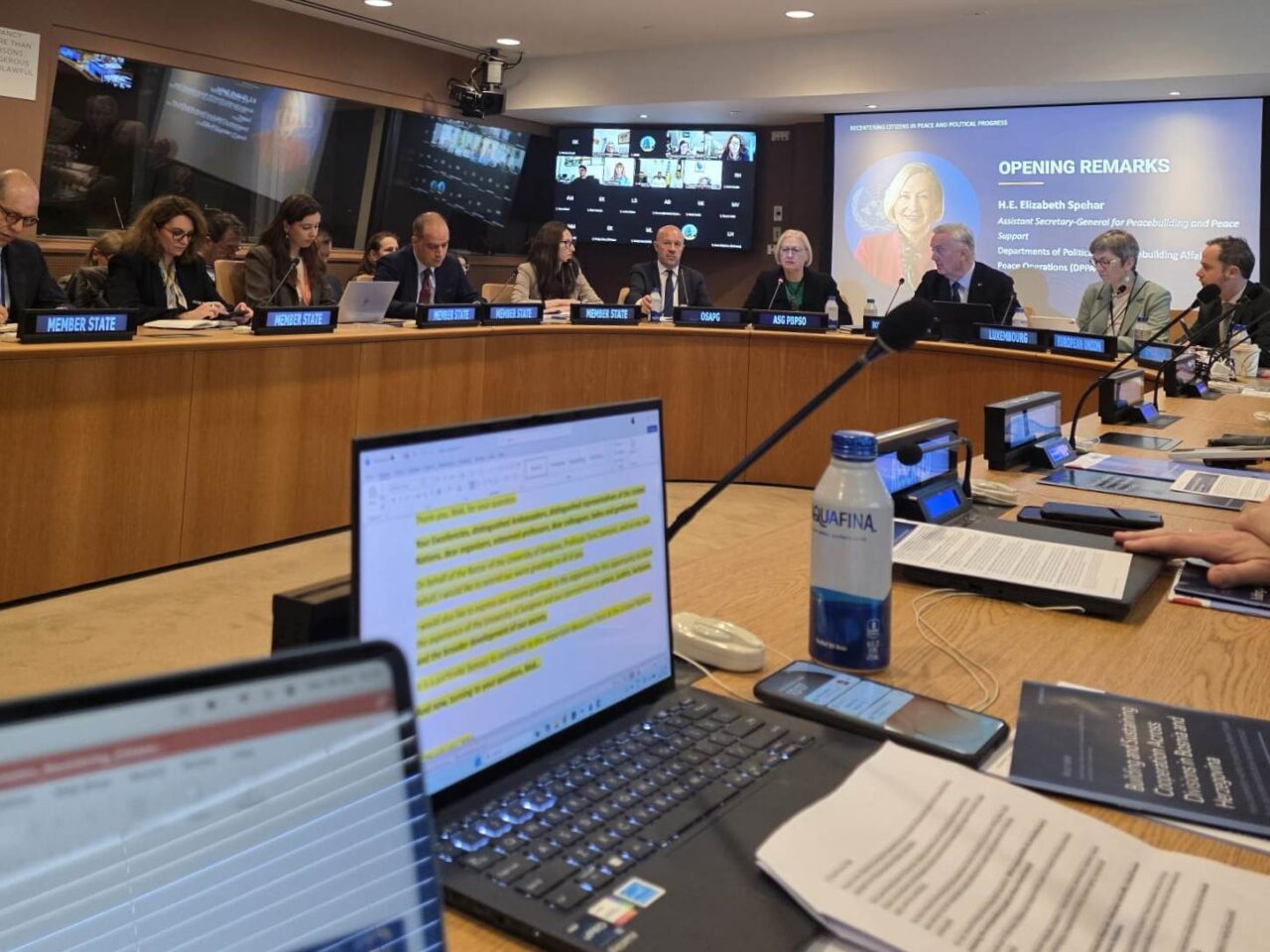 Participants attend the opening remarks, with remote attendees and a presentation displayed on screens.