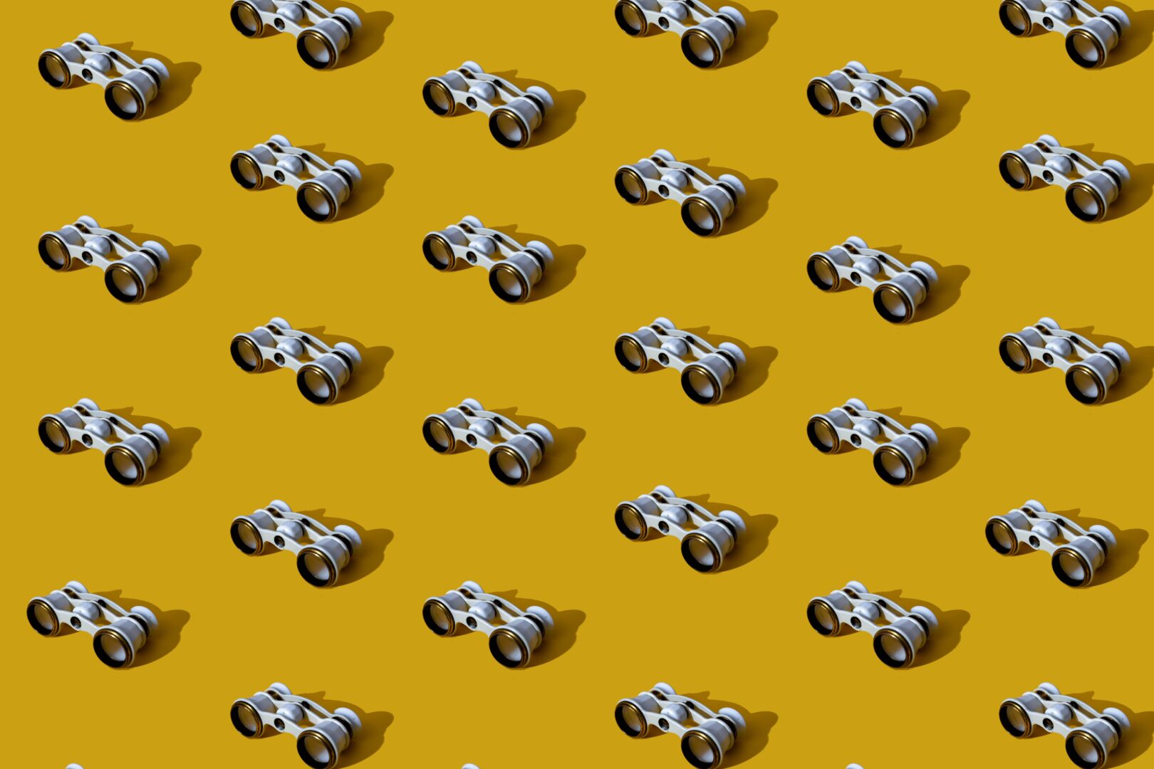 A repeating pattern of small white and silver opera glasses with long shadows on a mustard-yellow background.