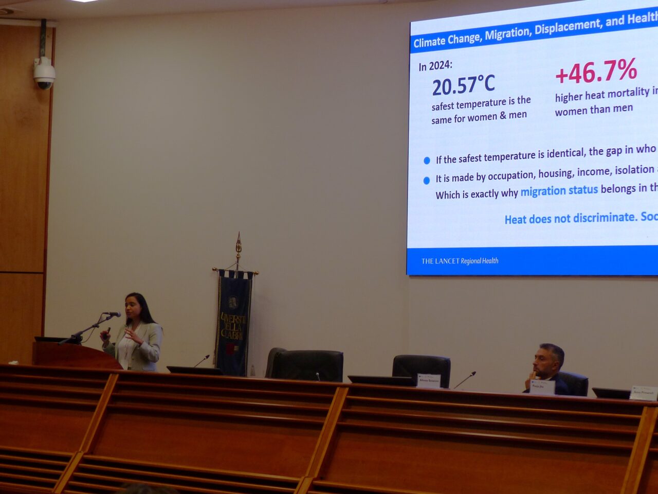 A woman in a light blazer speaks at a lectern with a microphone in a wood-panelled lecture hall. On the large screen beside her, a slide headed 'Climate Change, Migration, Displacement, and Health' states that in 2024 the safest temperature was 20.57°C for both women and men, and that heat mortality was 46.7% higher in women than in men. A man sits at the long desk in the foreground.