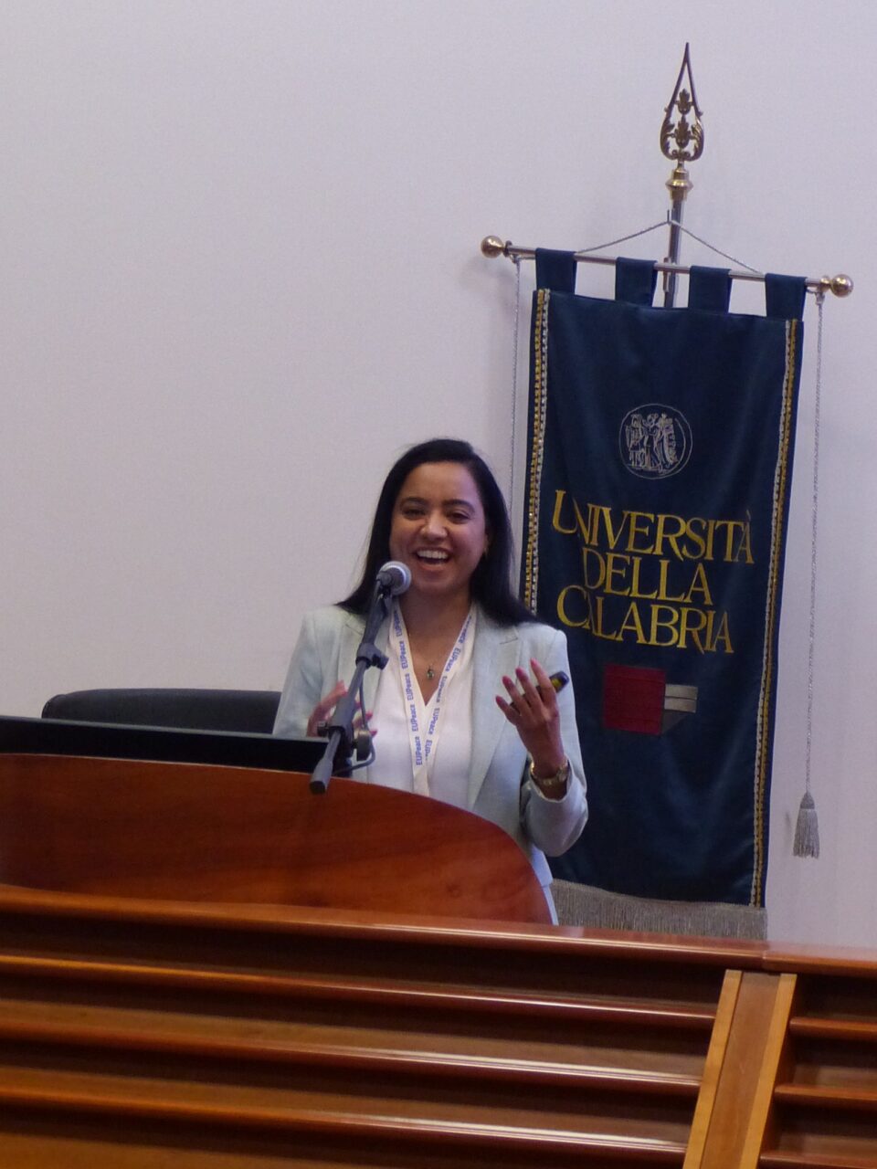 A smiling woman with long dark hair, wearing a light blazer, white blouse and an EUPeace lanyard, speaks into a microphone at a wooden lectern with her hands raised. Behind her hangs a dark blue banner reading "Università della Calabria".