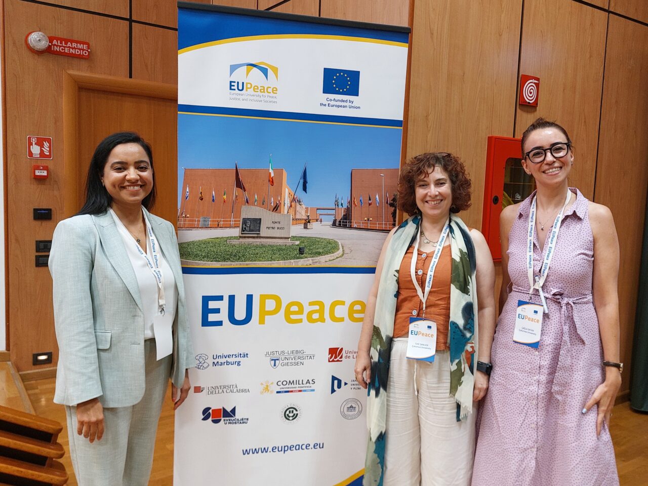 Three smiling women stand side by side in front of a EUPeace roll-up banner in a wood-panelled hall. The banner shows the EUPeace and European Union logos, a photograph of a building with flags, the website www.eupeace.eu and the logos of several partner universities. Two of the women wear EUPeace lanyards with name badges.
