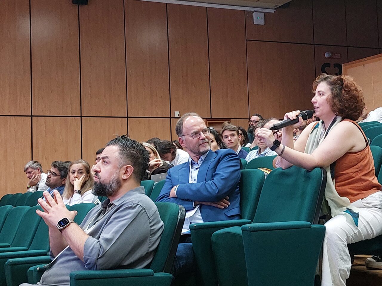 A woman with curly hair, seated on the edge of a green auditorium seat, speaks into a handheld microphone. In front of her, a man in a blue blazer and glasses listens with his arms crossed, and a bearded man in a grey shirt gestures with his hands. Other audience members sit behind them in a room with wood-panelled walls.