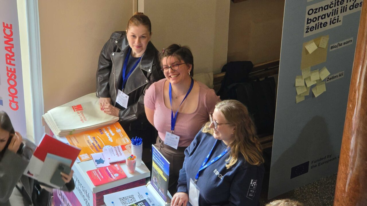 Three university representatives stand behind a University of Limoges information stand displaying brochures, promotional materials and university information.