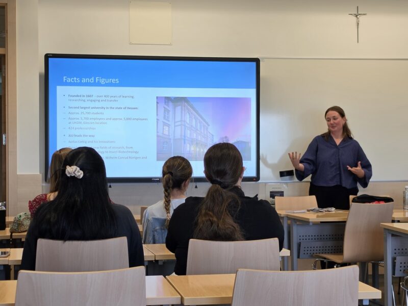 A woman gives a presentation at the front of a classroom while students sit facing a screen displaying a slide titled “Facts and Figures