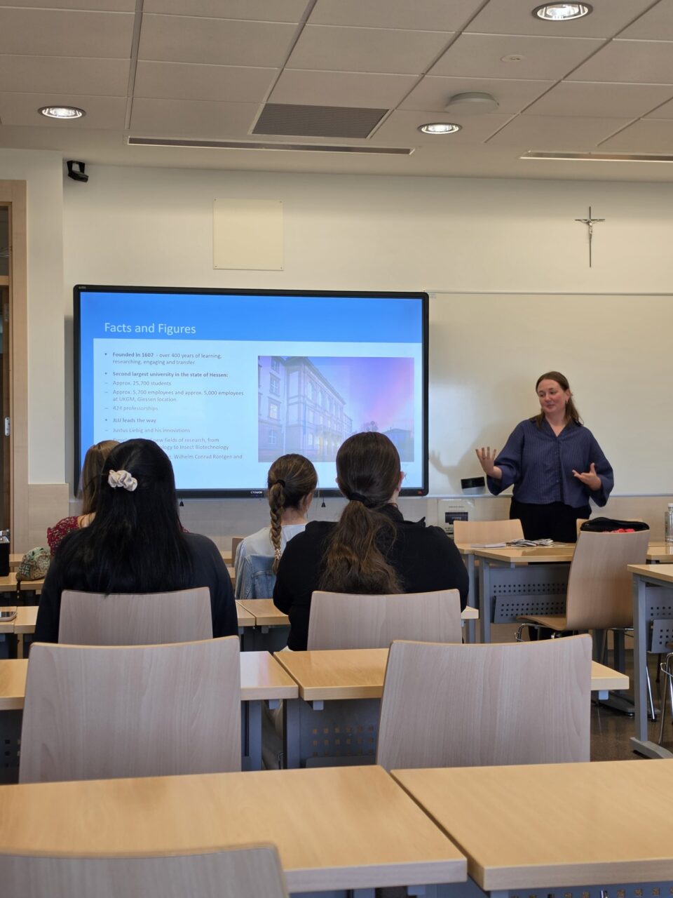 A woman gives a presentation at the front of a classroom while students sit facing a screen displaying a slide titled “Facts and Figures