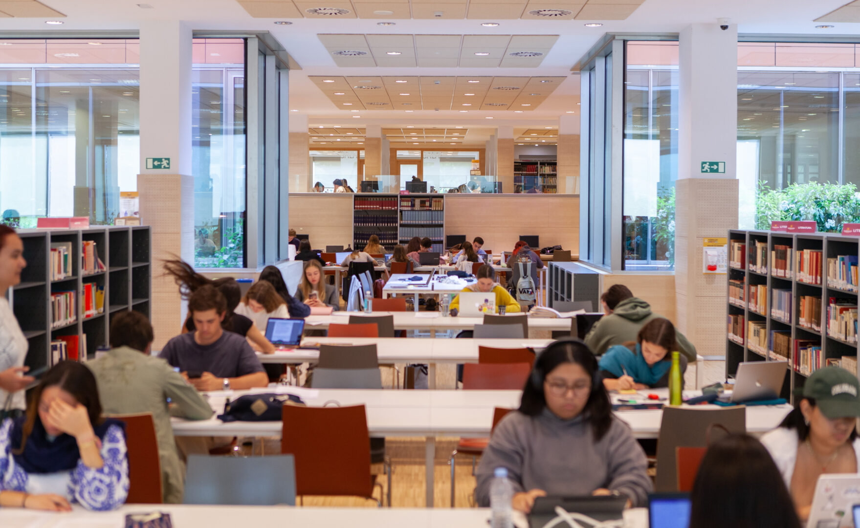 Interior of the Comillas Pontifical University Library showing tall wooden bookshelves, arched windows, and long study tables under warm lighting.