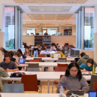 Interior of the Comillas Pontifical University Library showing tall wooden bookshelves, arched windows, and long study tables under warm lighting.