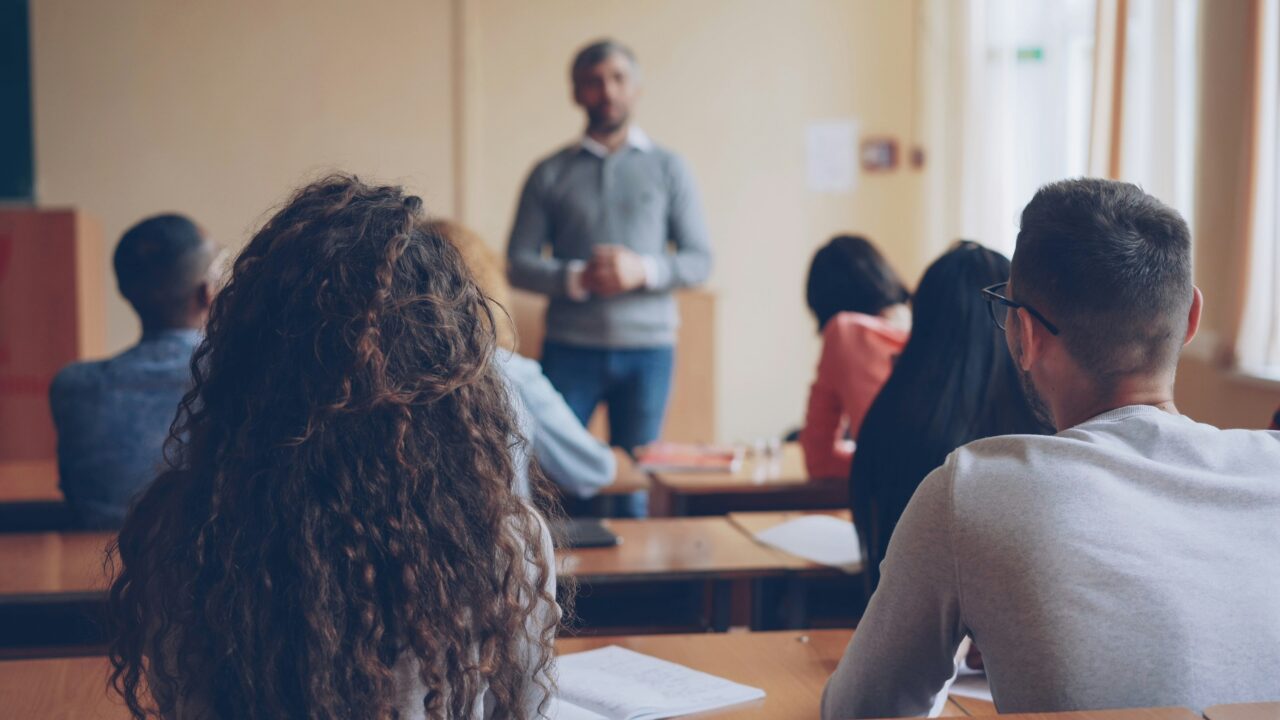 Students sitting in a classroom, photographed from behind. The teacher is standing in front of the class.