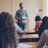Students sitting in a classroom, photographed from behind. The teacher is standing in front of the class.