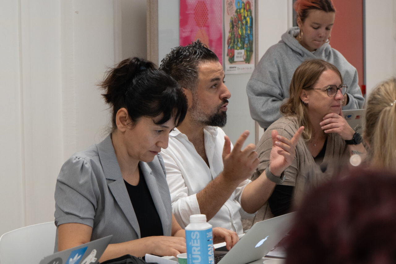 Participants sit at a table with laptops in a bright meeting room. A bearded man in a white shirt gestures with both hands as he speaks, while a woman in a grey blazer looks down beside him and a woman with glasses rests her chin on her hand and listens. Another woman in a grey hooded top stands behind them, and a blue water bottle is in the foreground.