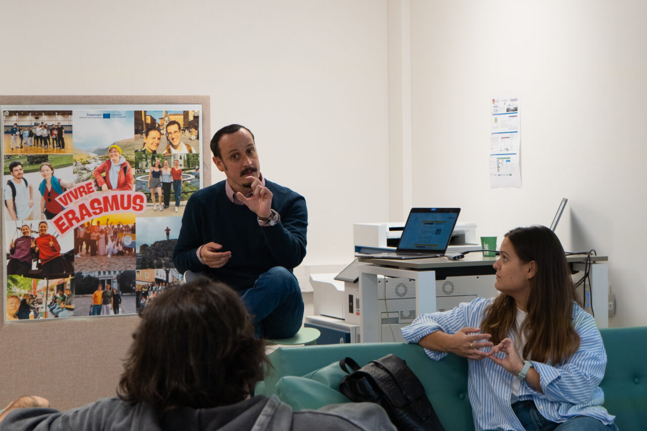 A man with a moustache, wearing a dark jumper and jeans, sits on a low stool and gestures with both hands while speaking to a woman with long brown hair in a striped blue shirt, who sits on a green sofa and looks towards him. The back of another person's head is in the foreground. Behind them hangs a collage of photographs of young people headed 'Vivre Erasmus', with an Erasmus+ logo. A laptop and a printer stand on a desk in the background.