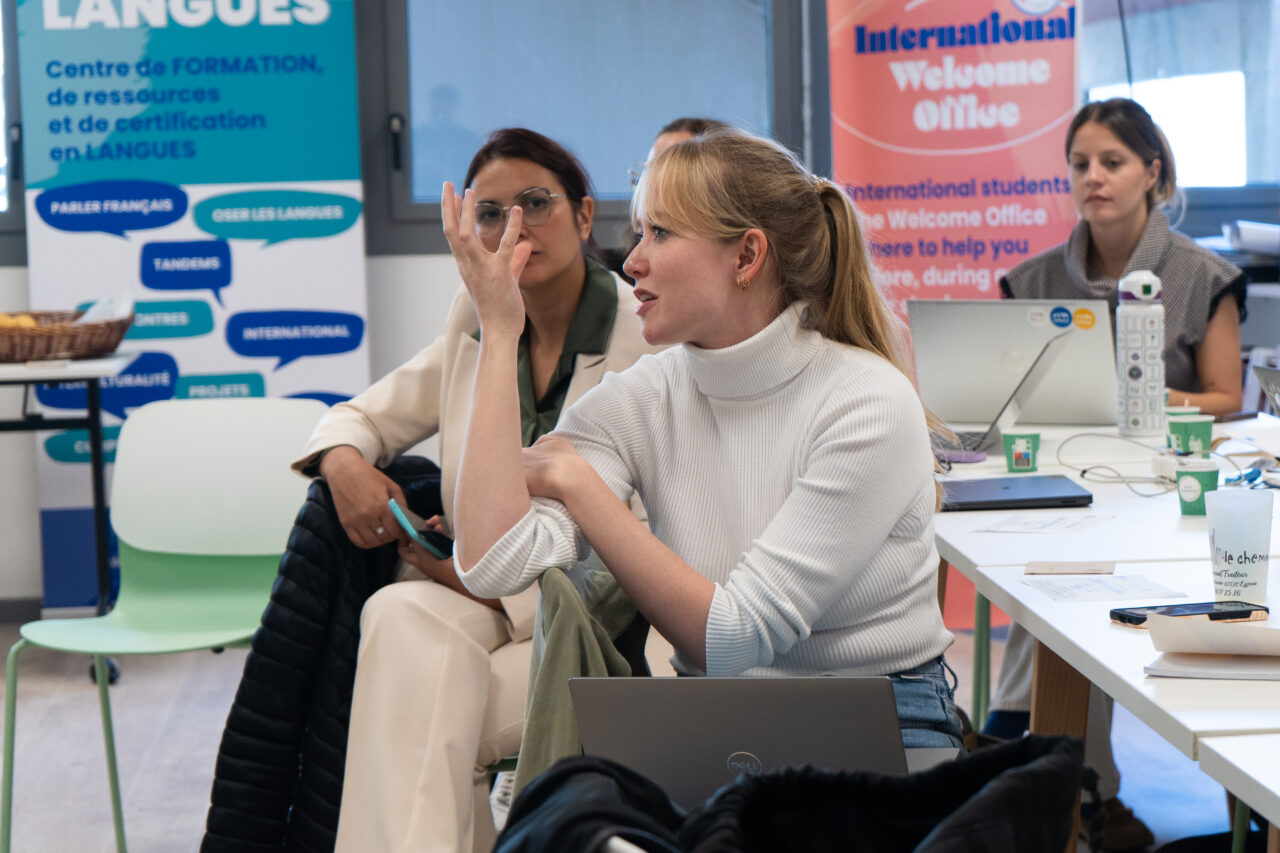 A woman with blonde hair in a ponytail and a white ribbed polo-neck jumper gestures with one raised hand as she speaks, seated at a table with a laptop. Behind her, a woman with glasses and a cream blazer and another woman in a grey top look on. Banners in the background read "Langues" with French text about language training, and "International Welcome Office".