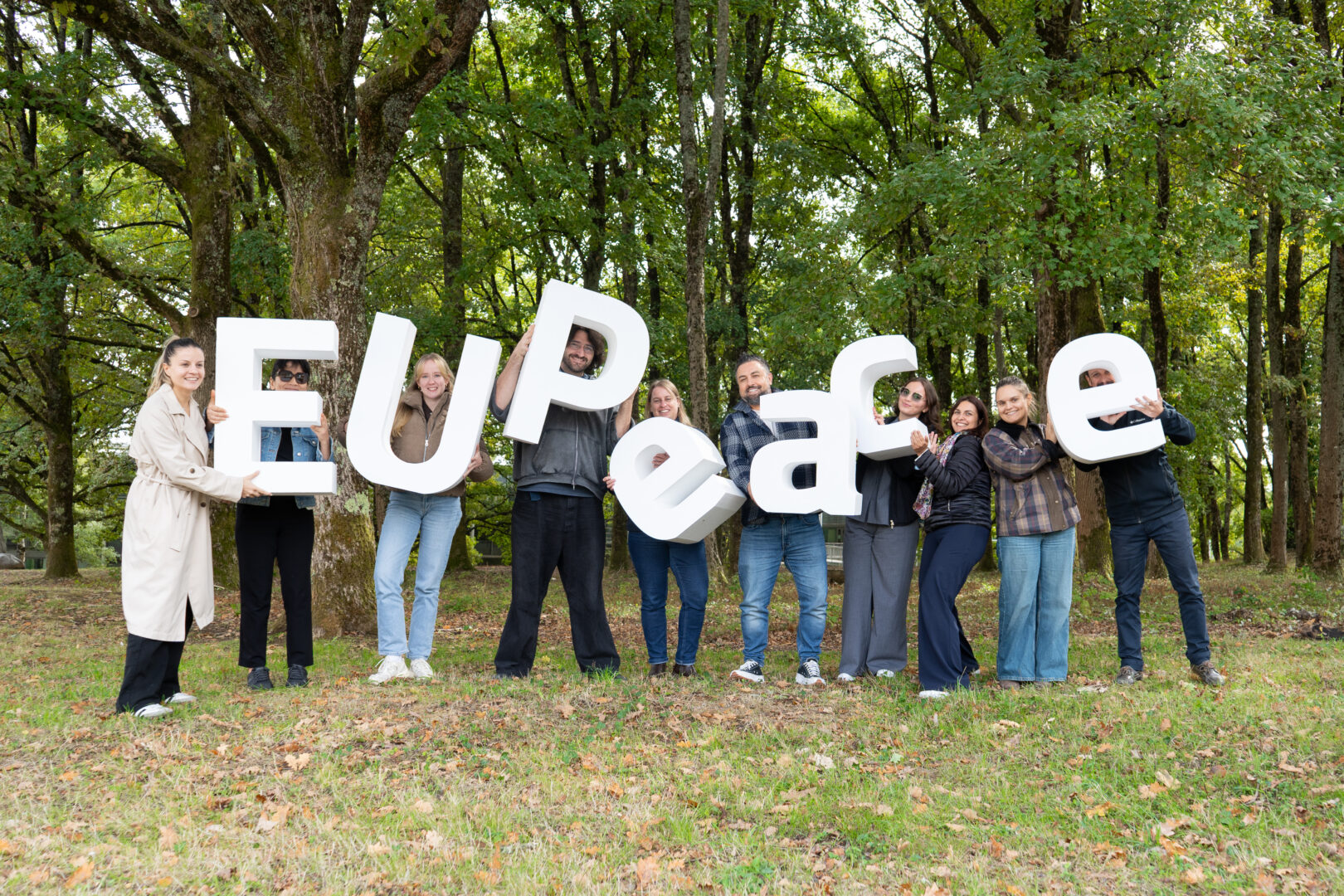 Ten people stand in a row on grass among tall trees, each holding up a large white three-dimensional letter. The letters are arranged to spell "EUPeace". Most of the group are smiling at the camera, and fallen leaves cover the ground.