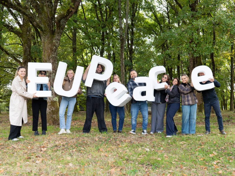 Ten people stand in a row on grass among tall trees, each holding up a large white three-dimensional letter. The letters are arranged to spell "EUPeace". Most of the group are smiling at the camera, and fallen leaves cover the ground.