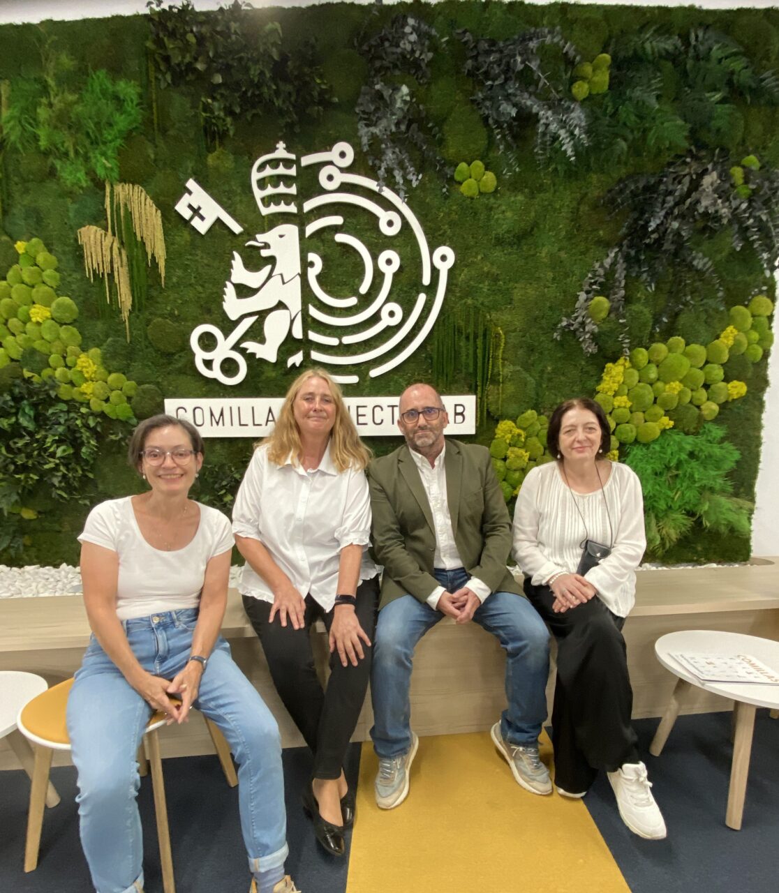 Four relaxed professors, three female, one male, sitting in front of Comillas' logo after debating their European Track Course "Exile and Migration in World Literature".