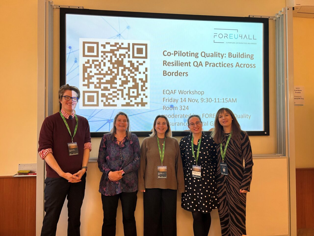 One man and four women posing in front of a projector wall for a group picture. The four persons are the workshop moderators for a workshop on Quality Assurance at the European Quality Assurance Forum 2025