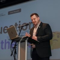The President of CAAI, Lukáš Benzl, standing in front of a large screen, giving his keynote address.