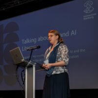 A blonde woman standing in front of a blue screen showing the title of her presentation "Language about AI and AI literacy - linguistic representations of generative AI and the implications for AI literacy in higher education"