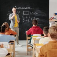 A teacher standing in front of a chalkboard asking his students a question.