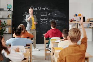 A teacher standing in front of a chalkboard asking his students a question.
