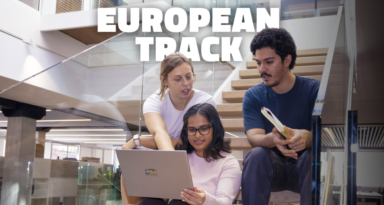 Three students sit together on a wooden staircase in a modern building with glass railings. In the middle, a student with glasses holds a laptop with the EUPeace logo on its lid, while a student behind her points at the screen and a student on the right holds a notebook. Large white letters at the top read "European Track".