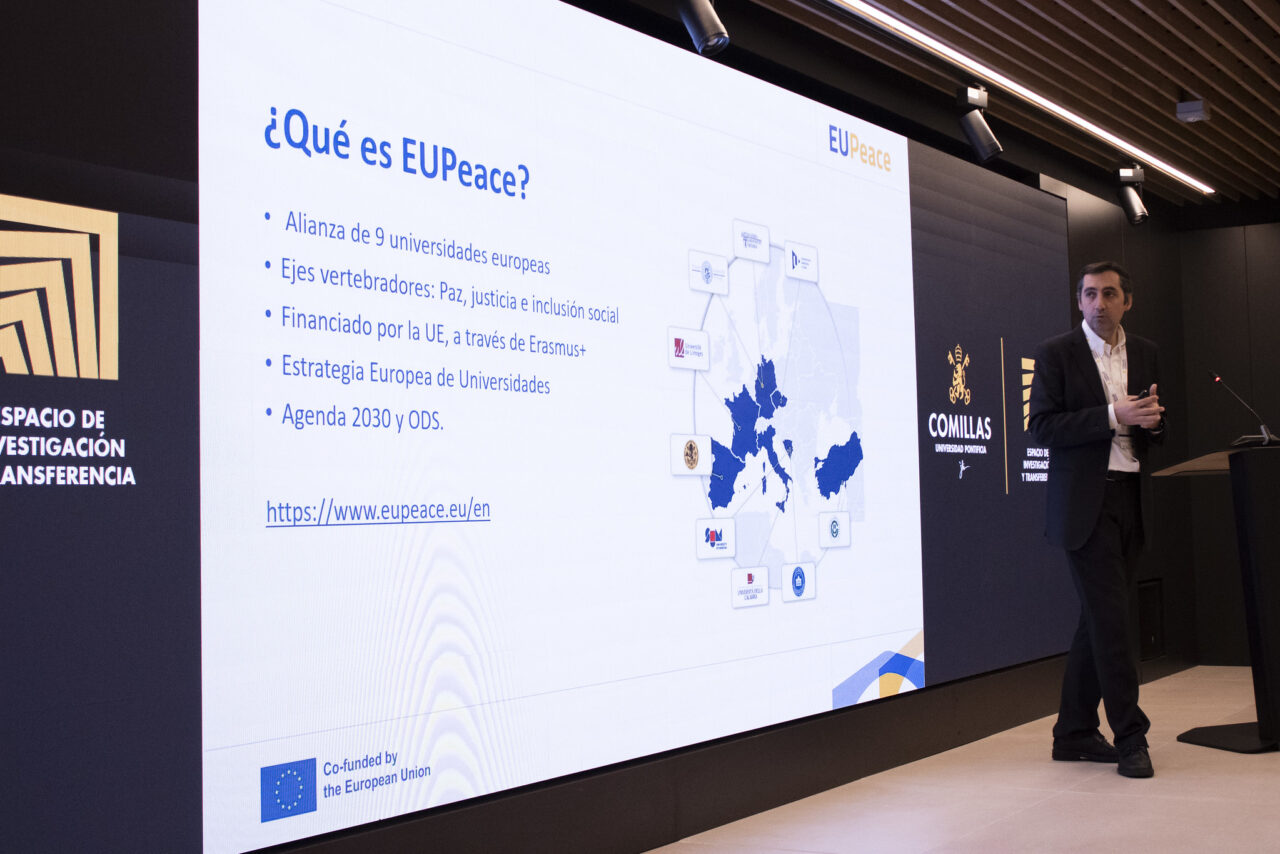 A presenter stands beside a large screen displaying information about the EUPeace alliance.
