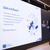 A presenter stands beside a large screen displaying information about the EUPeace alliance.