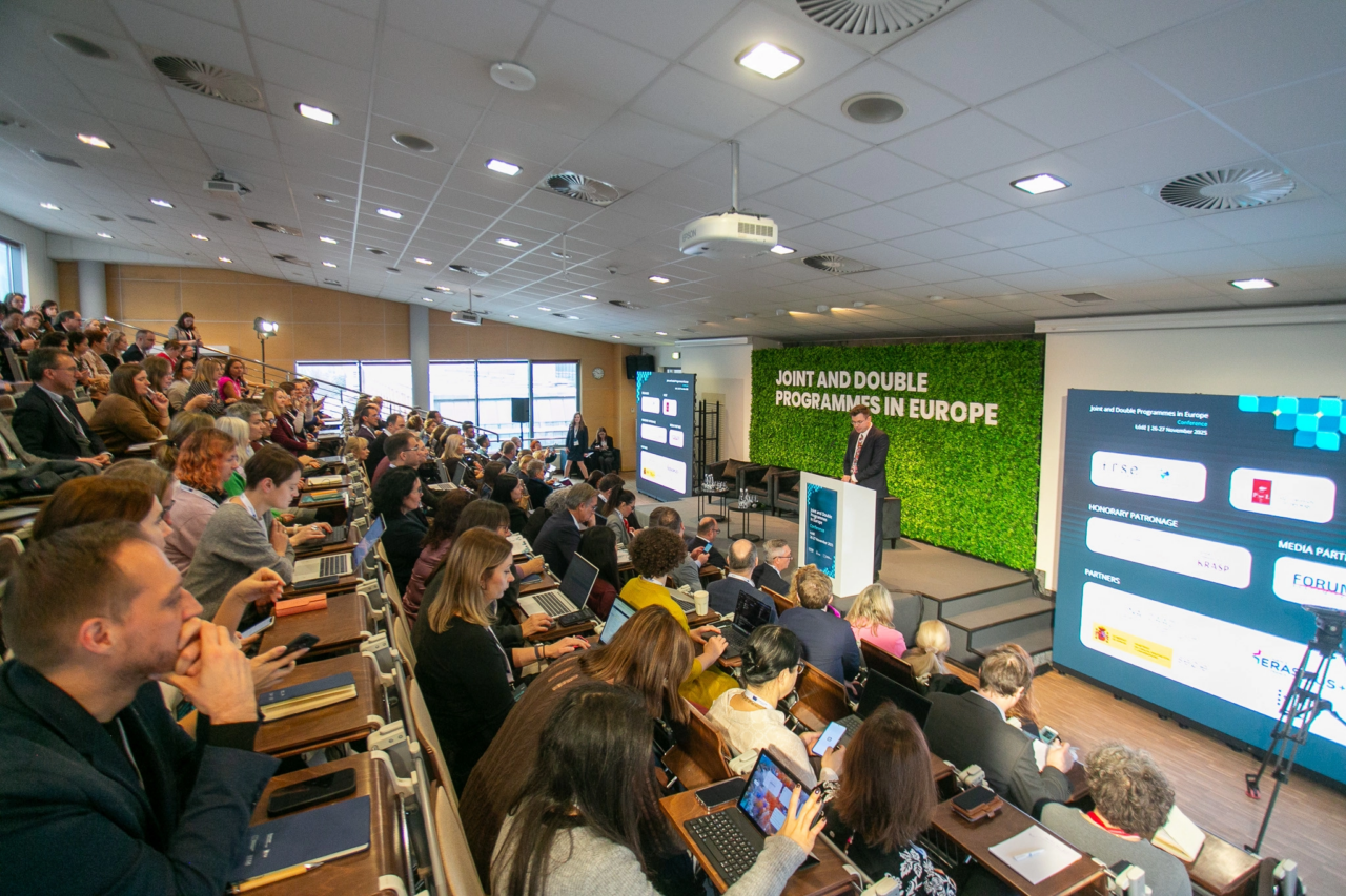 Several people sitting at tables in a conference room, looking at laptops and documents. A speaker is visible at the front.