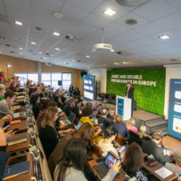 Several people sitting at tables in a conference room, looking at laptops and documents. A speaker is visible at the front.