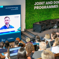 A panelist appearing on a screen via video call. Attendees listen attentively
