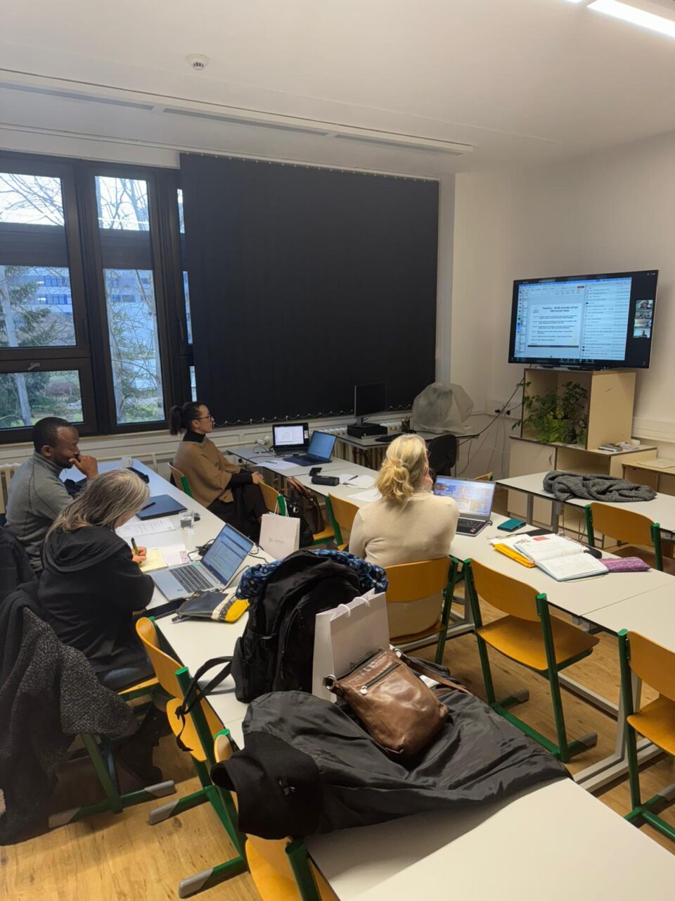 Four workshop participants with laptops seated at school desks, facing a wall-mounted screen with a presentation in a bright classroom.