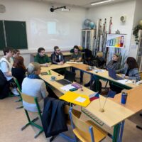 Small group discussion in a classroom setting, with students and a facilitator seated around tables.