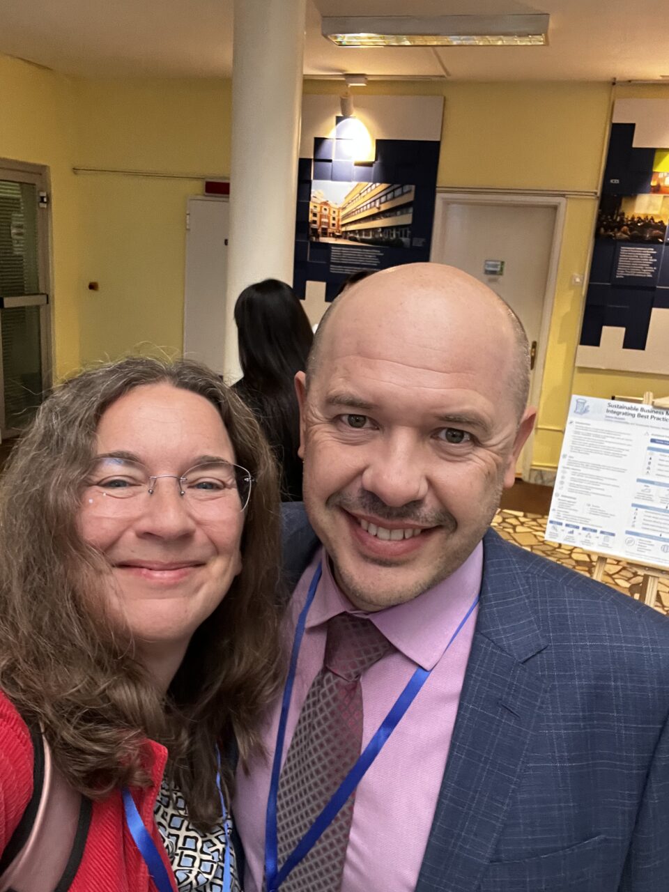 Two participants, Elisabeth Schulte and Adnan Efendić smile for a selfie in a university exhibition area, with a research poster behind them.