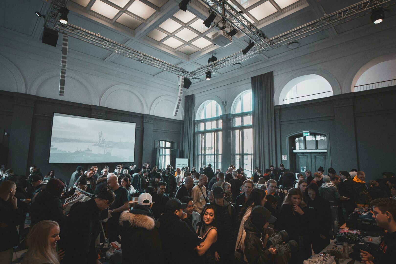 A large group of people networking in a spacious event hall with high arched windows and a projection screen in the background.