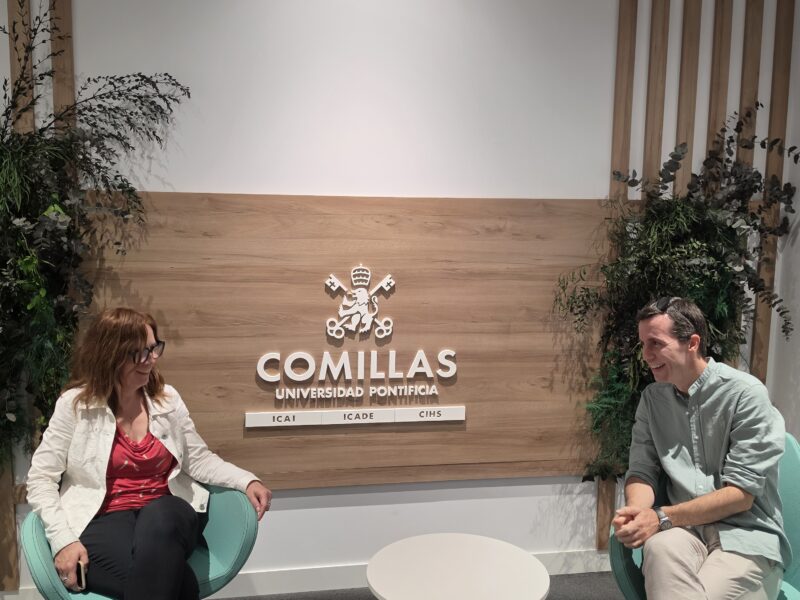 Two people sit facing each other in turquoise armchairs with a small round table between them in front of the Comillas Pontifical University sign, as if taking part in an interview.