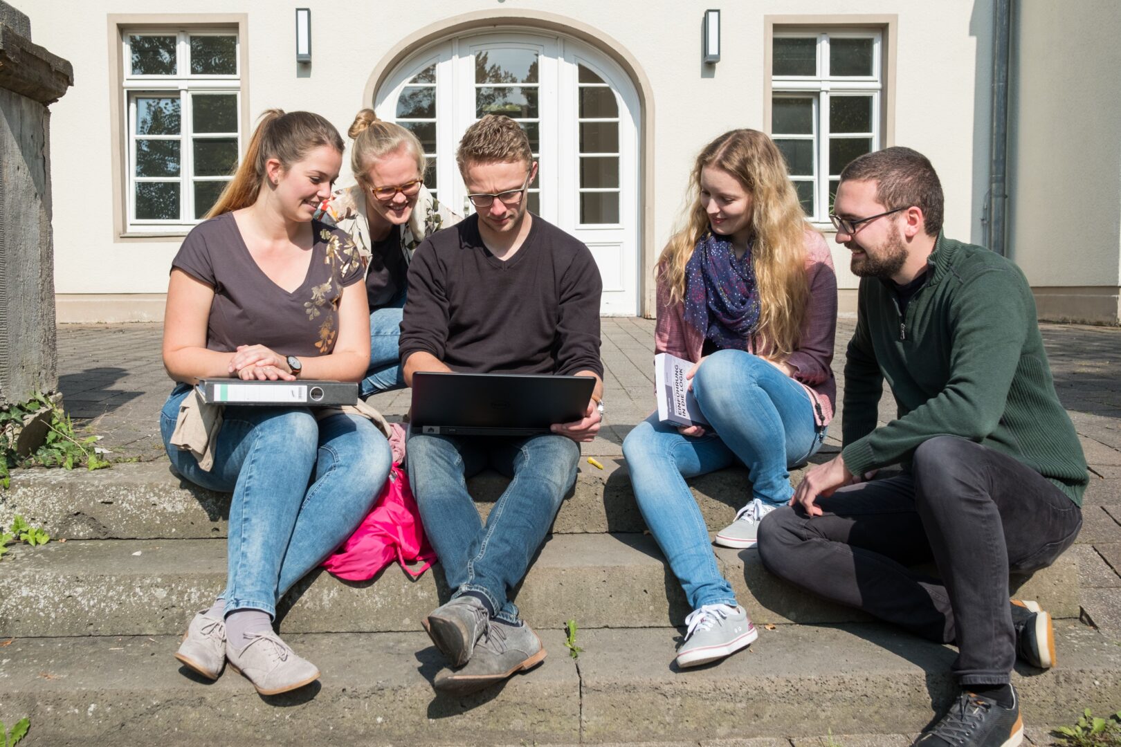 A group of students sitting on steps in front of a building entrance, looking at a laptop on the lap of one of the students.
