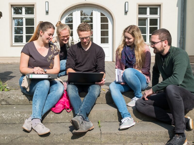 A group of students sitting on steps in front of a building entrance, looking at a laptop on the lap of one of the students.