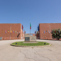 The Ponte Pietro Bucci at the University of Calabria campus in Rende, Italy. Two large terracotta-coloured buildings flank a central memorial stone, with international flags lining both sides under a clear blue sky.