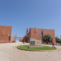 The Ponte Pietro Bucci at the University of Calabria campus in Rende, Italy, seen from a slight angle. A person walks past the central memorial stone. International flags line both sides of the bridge between the two terracotta-coloured buildings.