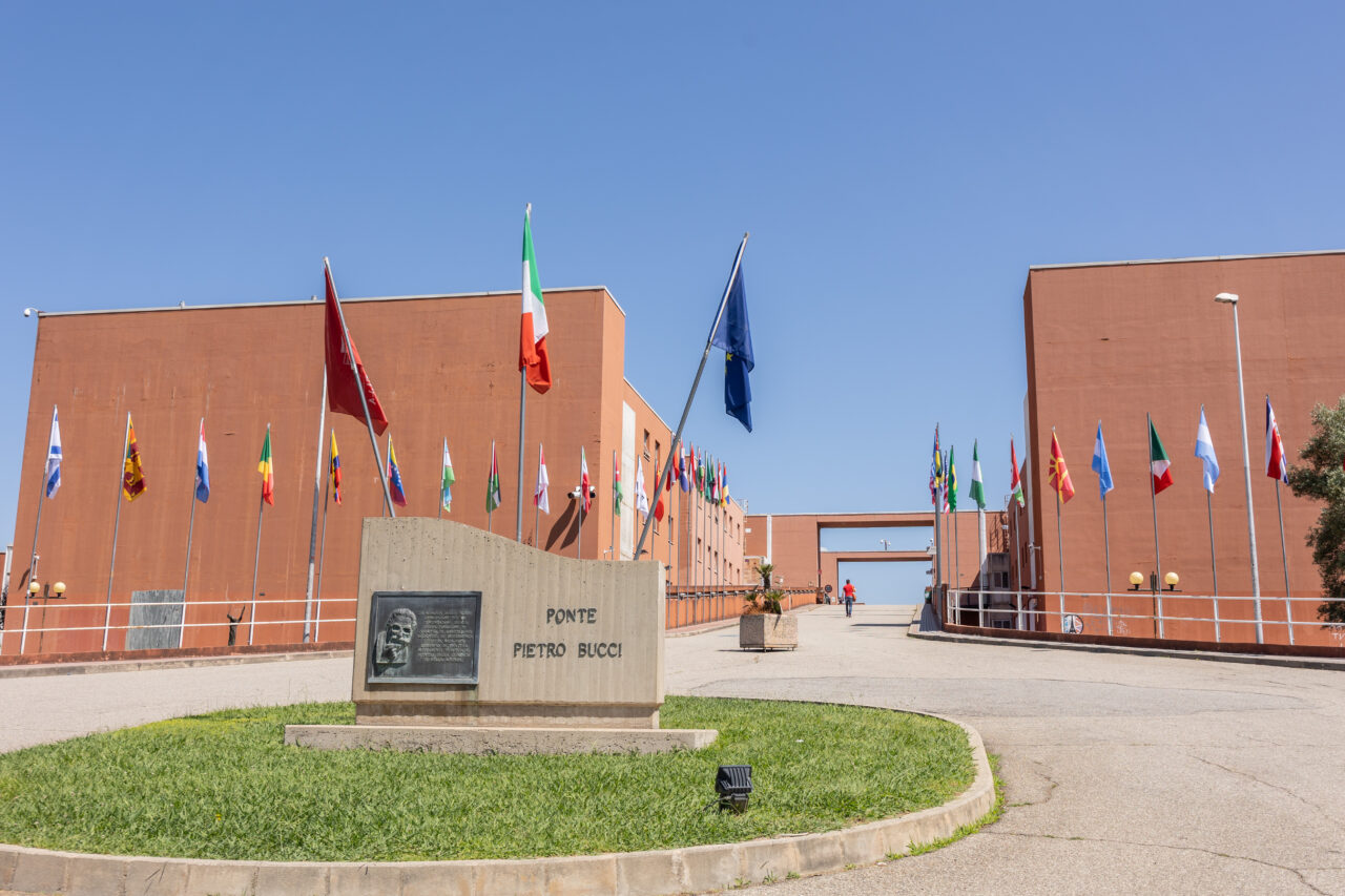 Close-up view of the Ponte Pietro Bucci memorial stone at the University of Calabria campus in Rende, with the Italian and European Union flags prominently visible. International flags and the bridge walkway extend into the background between the campus buildings.