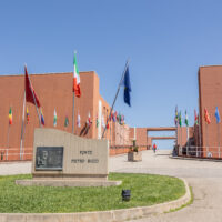 Close-up view of the Ponte Pietro Bucci memorial stone at the University of Calabria campus in Rende, with the Italian and European Union flags prominently visible. International flags and the bridge walkway extend into the background between the campus buildings.