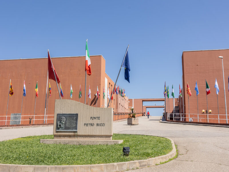Close-up view of the Ponte Pietro Bucci memorial stone at the University of Calabria campus in Rende, with the Italian and European Union flags prominently visible. International flags and the bridge walkway extend into the background between the campus buildings.