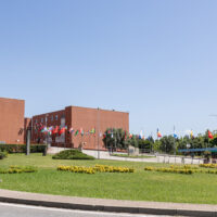 Wide view of the University of Calabria campus in Rende, showing the main buildings surrounded by green lawns, flower beds and international flags along the campus road.
