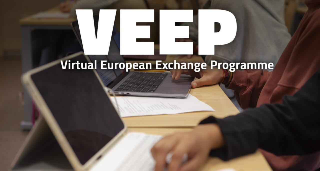 Close-up of several students working side by side at a wooden desk: hands on a laptop keyboard and trackpad, a tablet with a keyboard in the foreground and printed notes on the table. Large white letters at the top read "VEEP" with the subtitle "Virtual European Exchange Programme".
