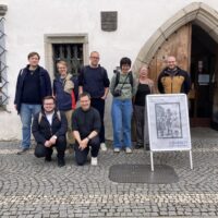 Group of nine people standing in front of the entrance of the Museum of Šumava in Sušice, with an exhibition sign reading “80x Díky!” placed in front.