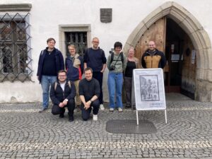 Group of nine people standing in front of the entrance of the Museum of Šumava in Sušice, with an exhibition sign reading “80x Díky!” placed in front.